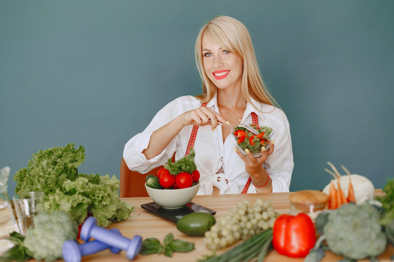 who-we-are Blonde woman enjoying a fresh salad surrounded by vegetables, promoting healthy eating.