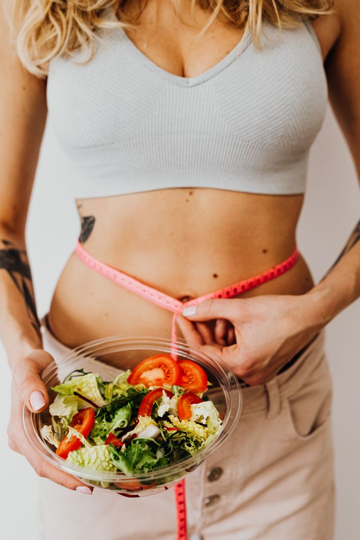 services-04 Fit woman in a crop top holds a salad bowl while measuring her waist, emphasizing healthy lifestyle and weight loss.