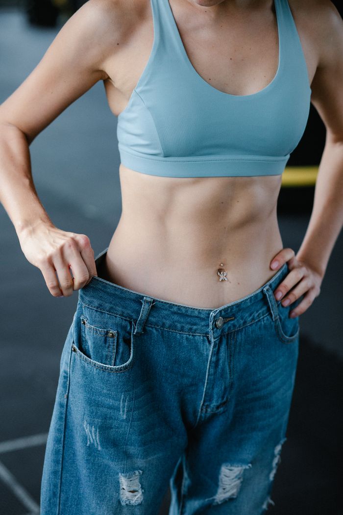 services-03 Fit woman shows weight loss in oversized jeans, displaying toned abs.