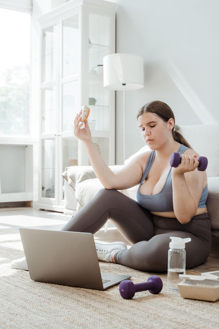 services-02 A plus size woman at home balancing fitness with lifestyle, using weights and enjoying a donut.